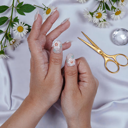 Hand with transparent press on nails featuring white floral design, surrounded by daisies and gold scissors on silk