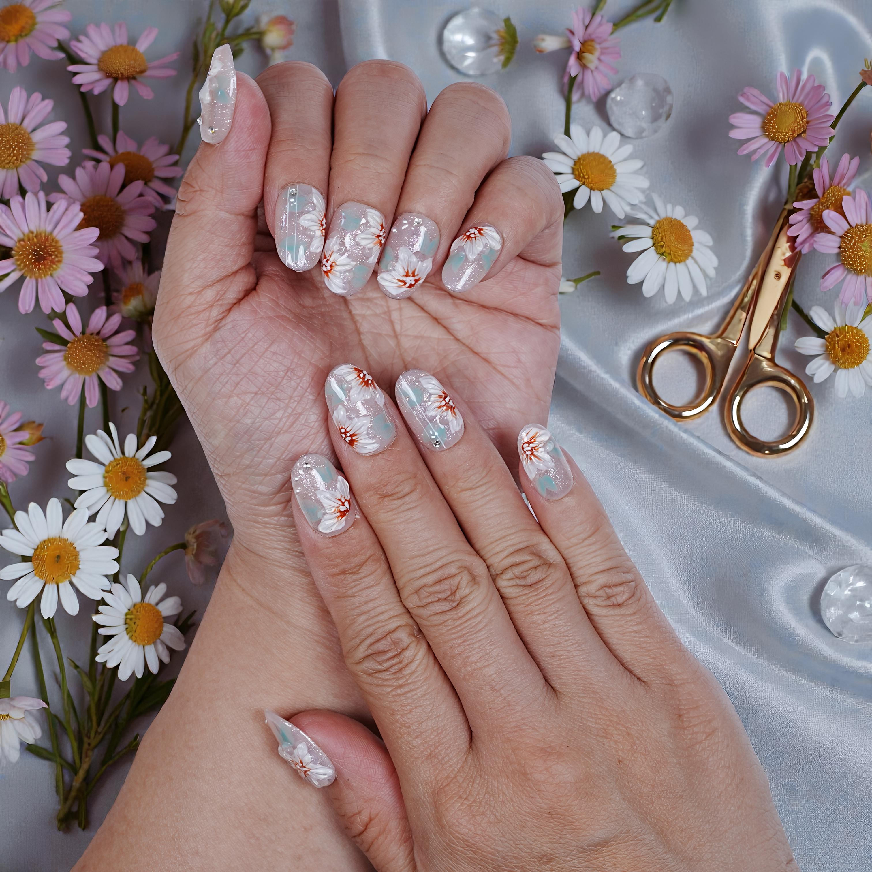 Hands with handmade press on nails featuring white floral designs on a glittery background, surrounded by fresh daisies and golden nail scissors on satin
