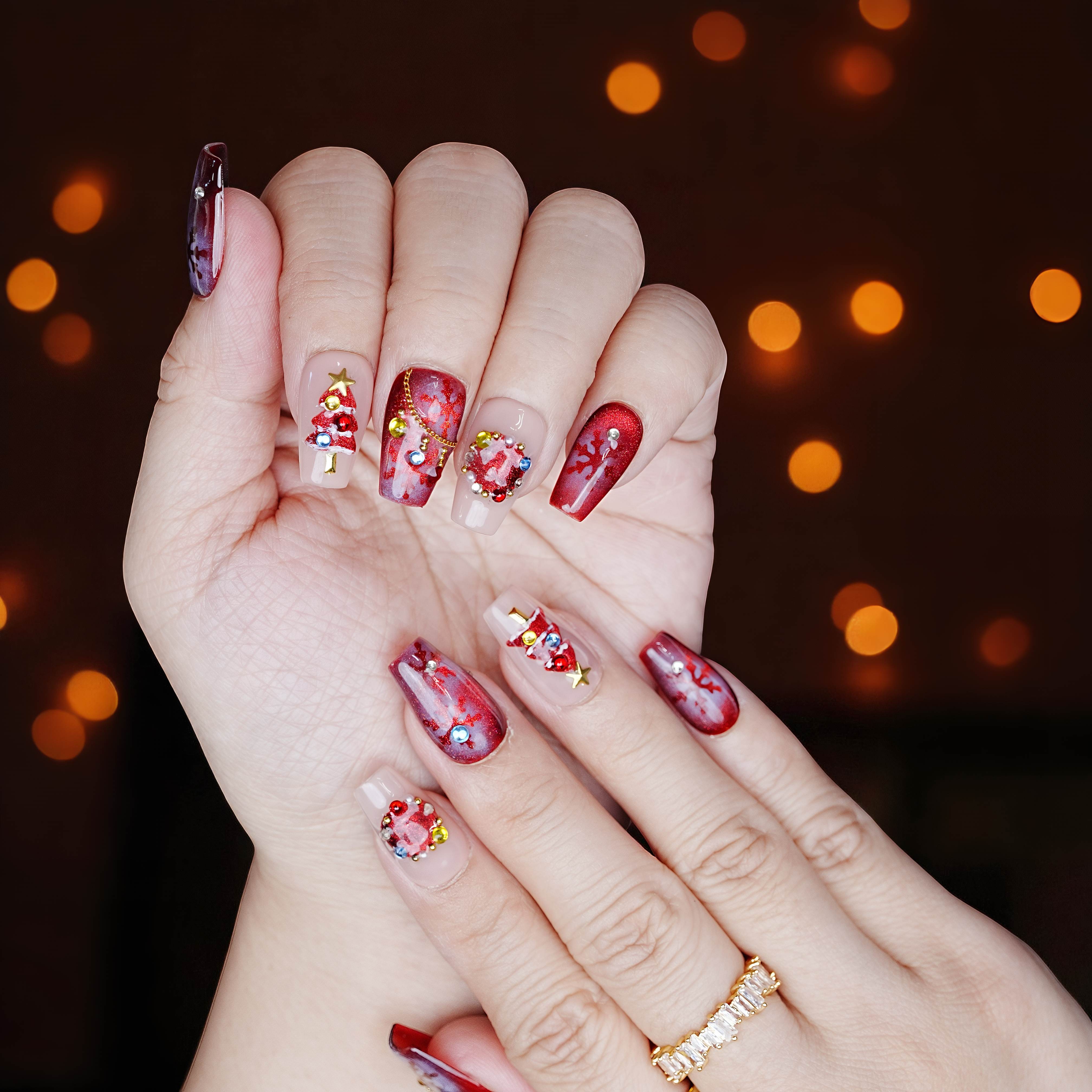 Holiday themed nails in festive red with 3D decorations, hand pose near golden lights