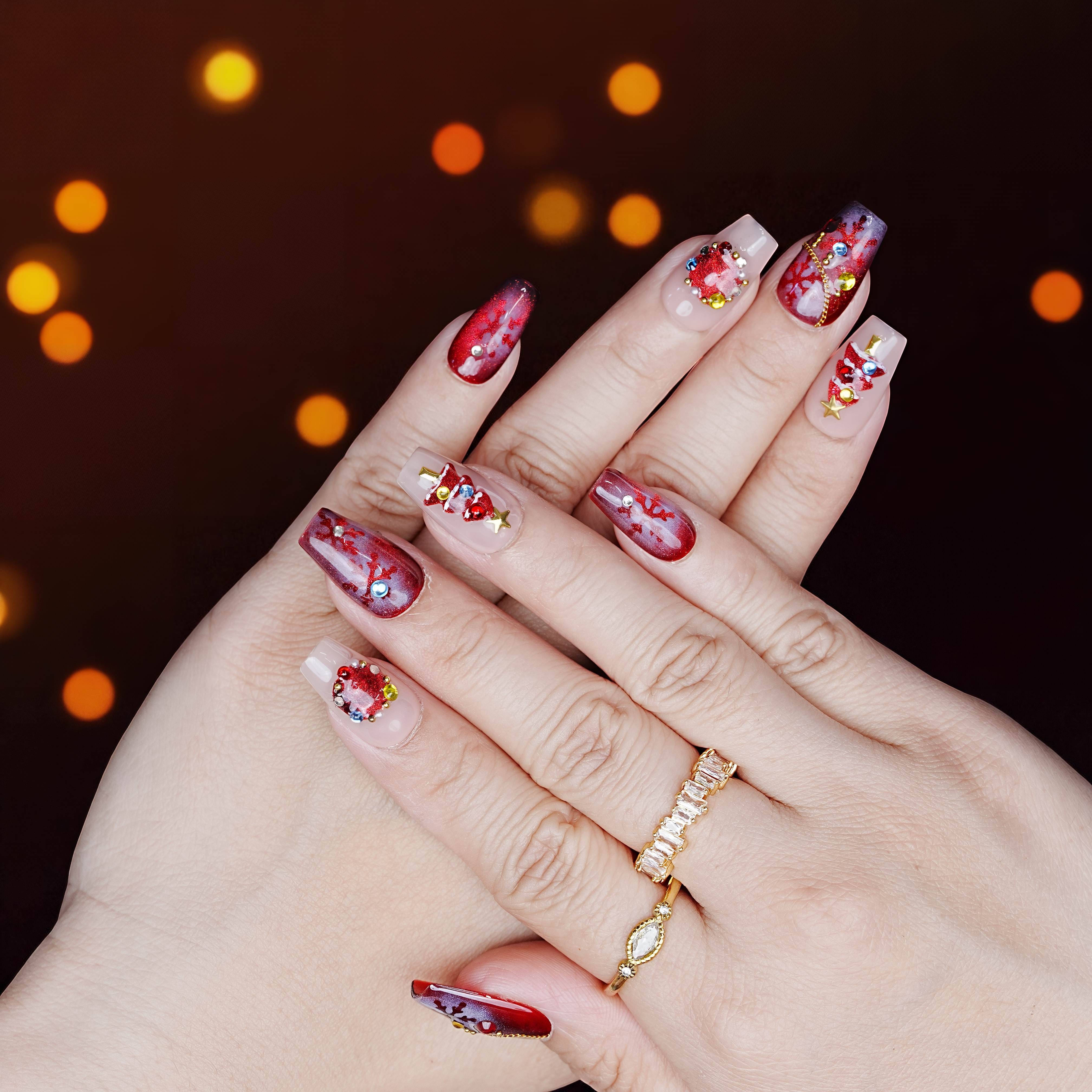 Red and nude press on nails with Christmas tree art and festive charms, hands posed on dark background with glowing bokeh