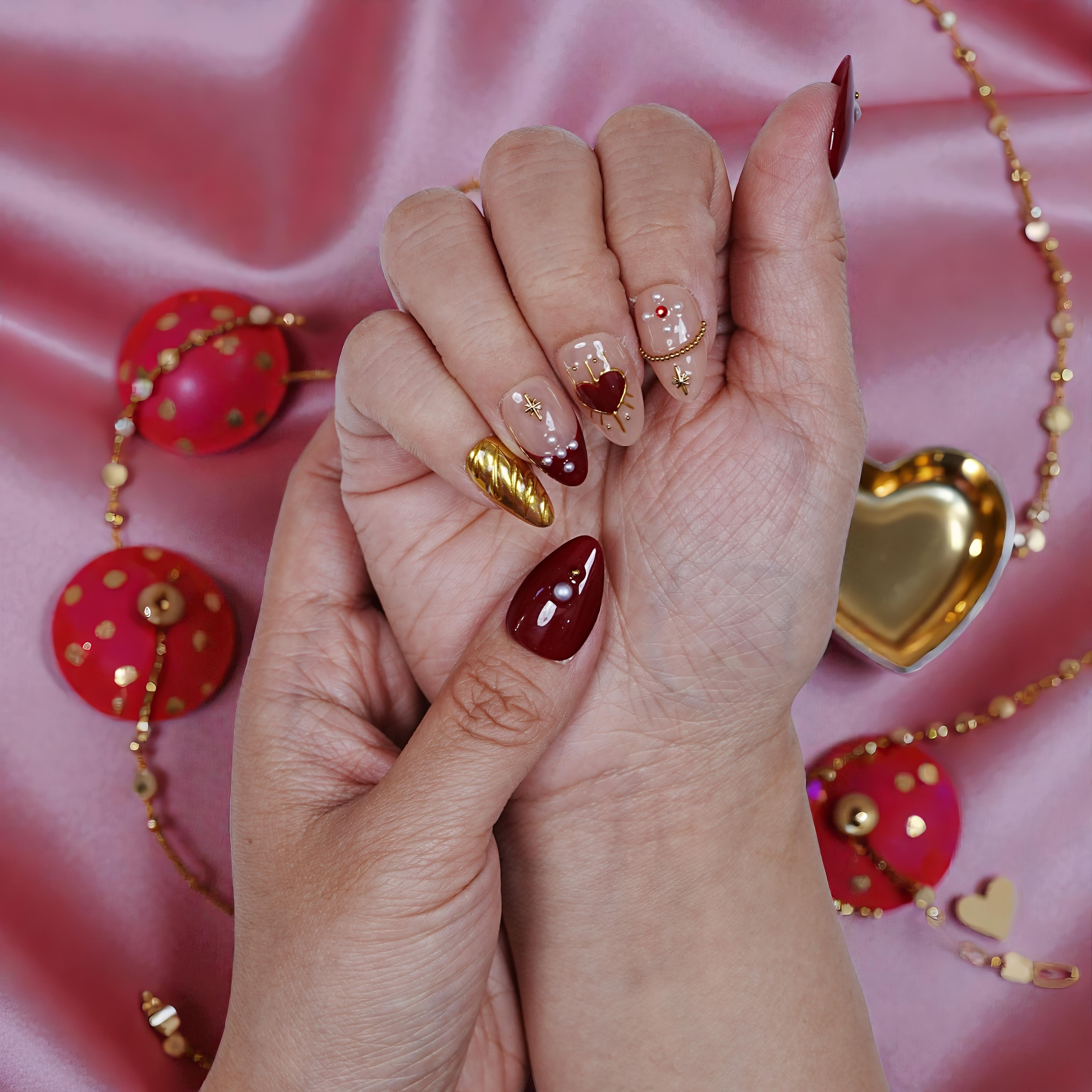 Hand with premium handmade press on nails in red, gold, and nude heart designs on pink satin.