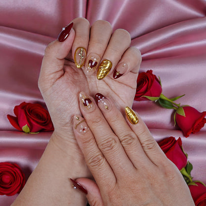 Handmade press on nails in burgundy and gold with heart accents, displayed on pink satin with roses.