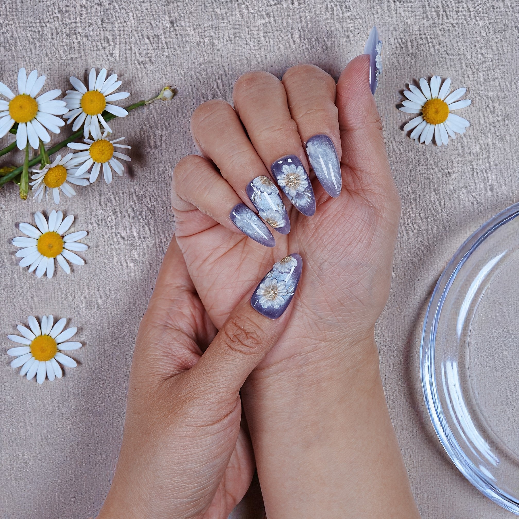 Hand with purple daisy floral press on nails, surrounded by daisies on a neutral fabric.