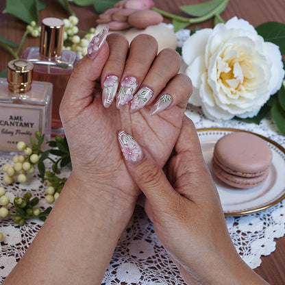 Hand with premium handmade press on nails featuring pink floral design, surrounded by perfume, flowers, and a macaron on a lace tablecloth.
