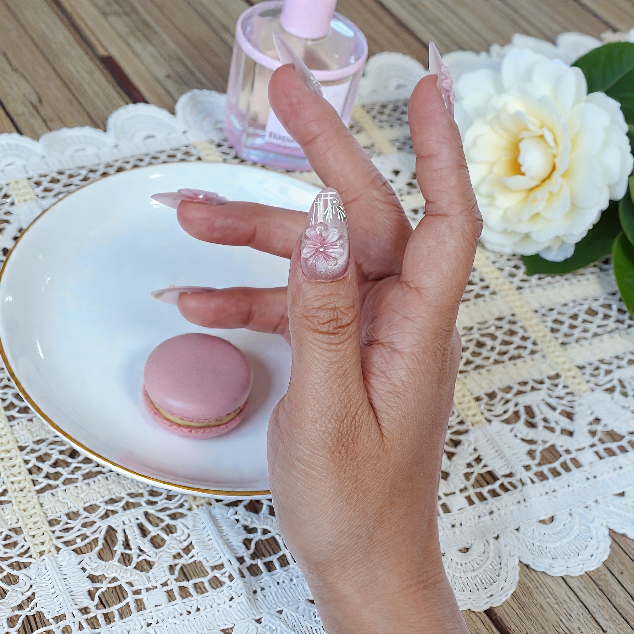 Hand with premium handmade floral press on nails holding macaron near perfume and flower